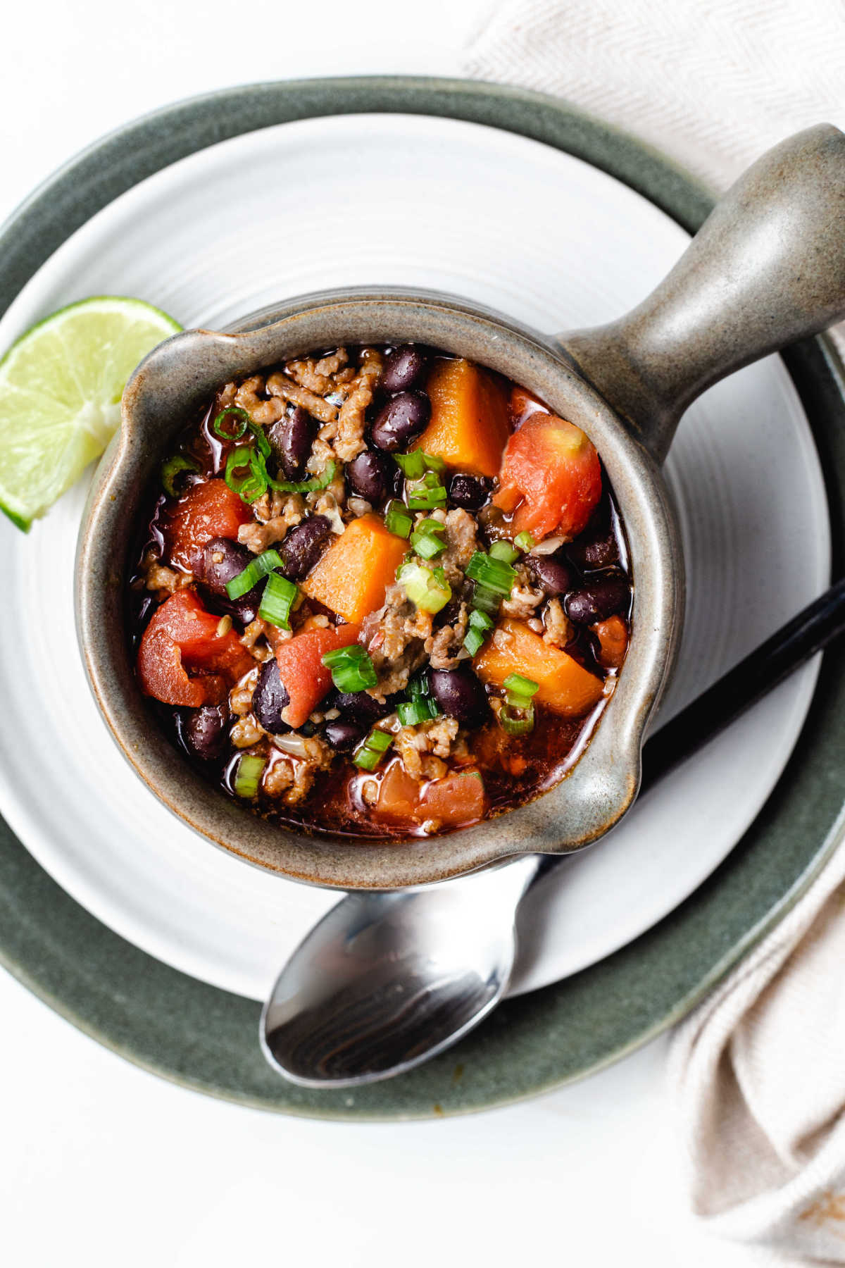top down view of a soup mug filled with black bean and sweet potato chili sitting on a white plate with a spoon laying to the side.