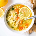 lemon chicken orzo soup in a white bowl with a slice of lemon and a spoon on a table.