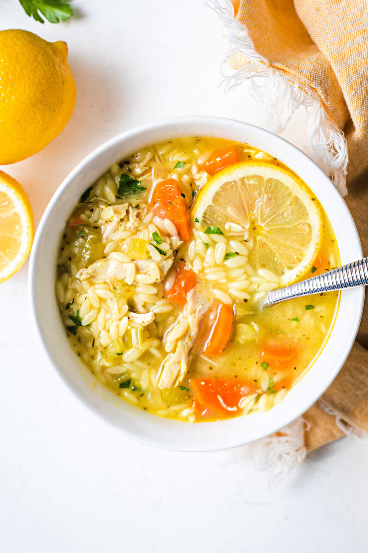 lemon chicken orzo soup in a white bowl with a slice of lemon and a spoon on a table.