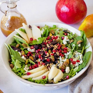 a white bowl with Christmas Salad on a table filled with greens, sliced pears, pomegranate arils, and pistachios.