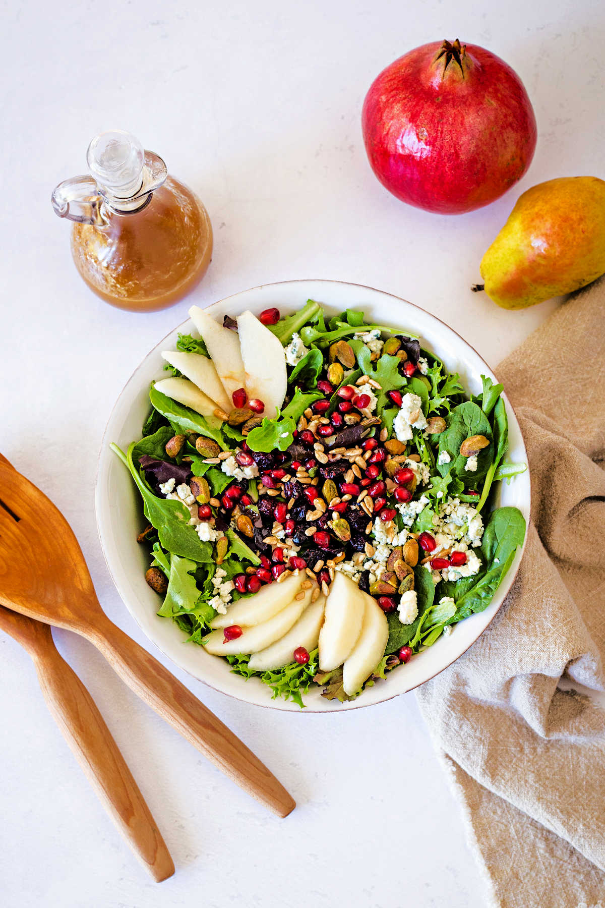 top down view of a white bowl with Christmas Salad on a table filled with greens, sliced pears, pomegranate arils, and pistachios with a curet of vinaigrette and a pomegranate sitting on a table.