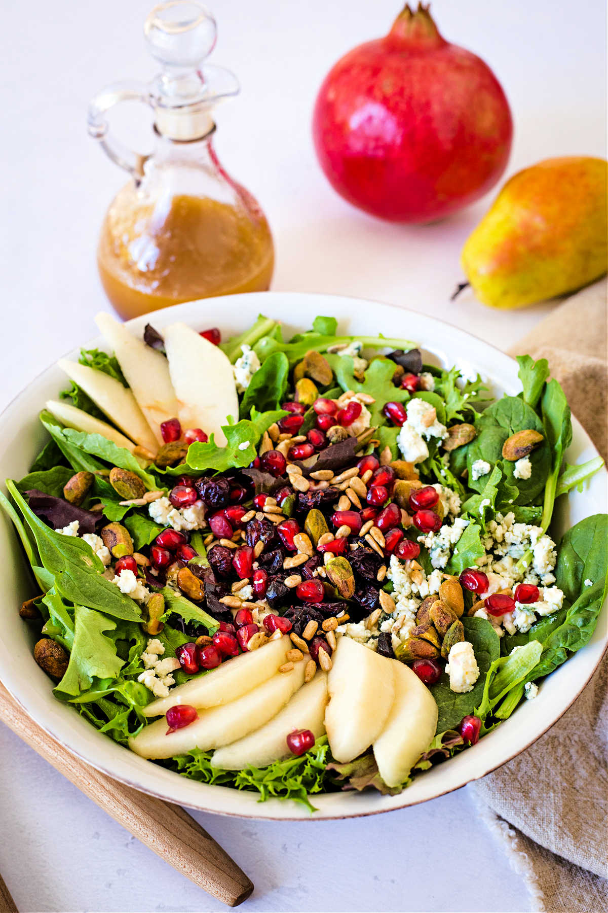 top down view of a white bowl with Christmas Salad on a table filled with greens, sliced pears, pomegranate arils, and pistachios with a curet of vinaigrette and a pomegranate sitting on a table.