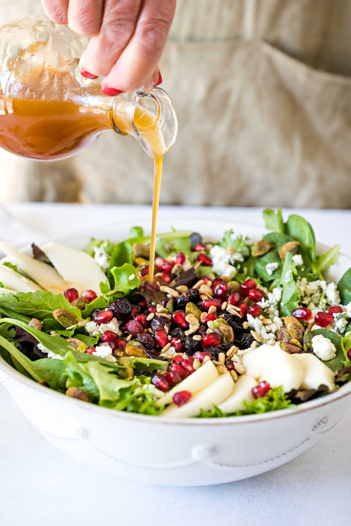 pouring cranberry maple vinaigrette over a bowl of salad greens with pears and pistachios.