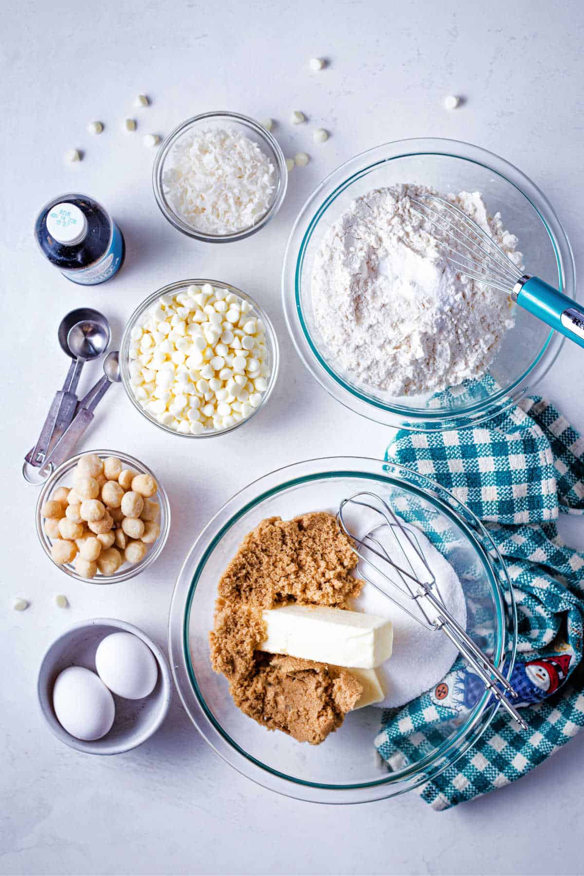 ingredients for white chocolate macadamia nut cookies on a table: butter, brown sugar, granulated sugar, eggs, flour, white chocolate chips, coconut, and macadamia nuts.