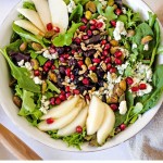 top down view of a white bowl with Christmas Salad on a table filled with greens, sliced pears, pomegranate arils, and pistachios with a curet of vinaigrette and a pomegranate sitting on a table.