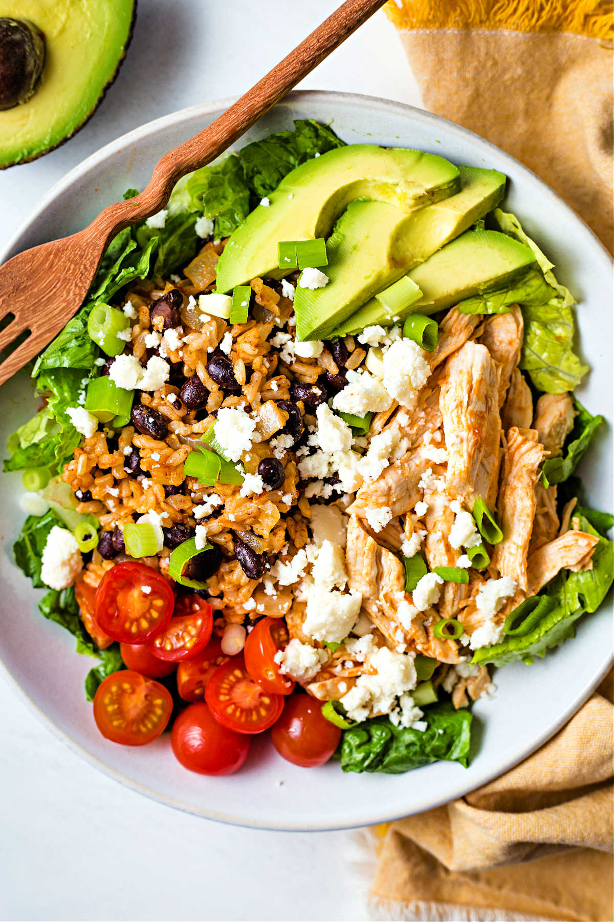 a bowl filled with lettuce, rice and beans, chicken, tomatoes, and avocado slices on a table.