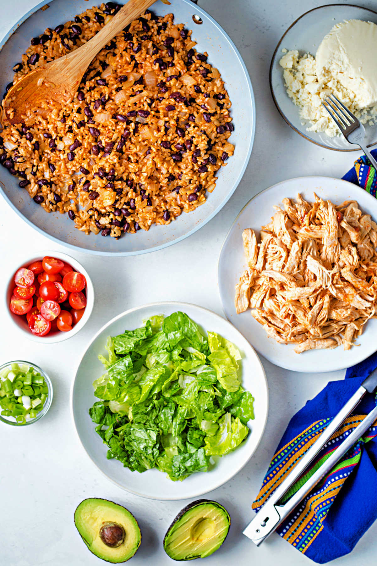 ingredients for building chicken burrito bowls on a table: rice and beans; shredded chicken; lettuce; tomatoes; avocado; queso fresca cheese.