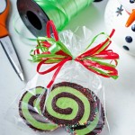 Christma pinwheel cookies packaged in a cellophane bag tied with red and green ribbon on a table with a spool of ribbon and scissors.