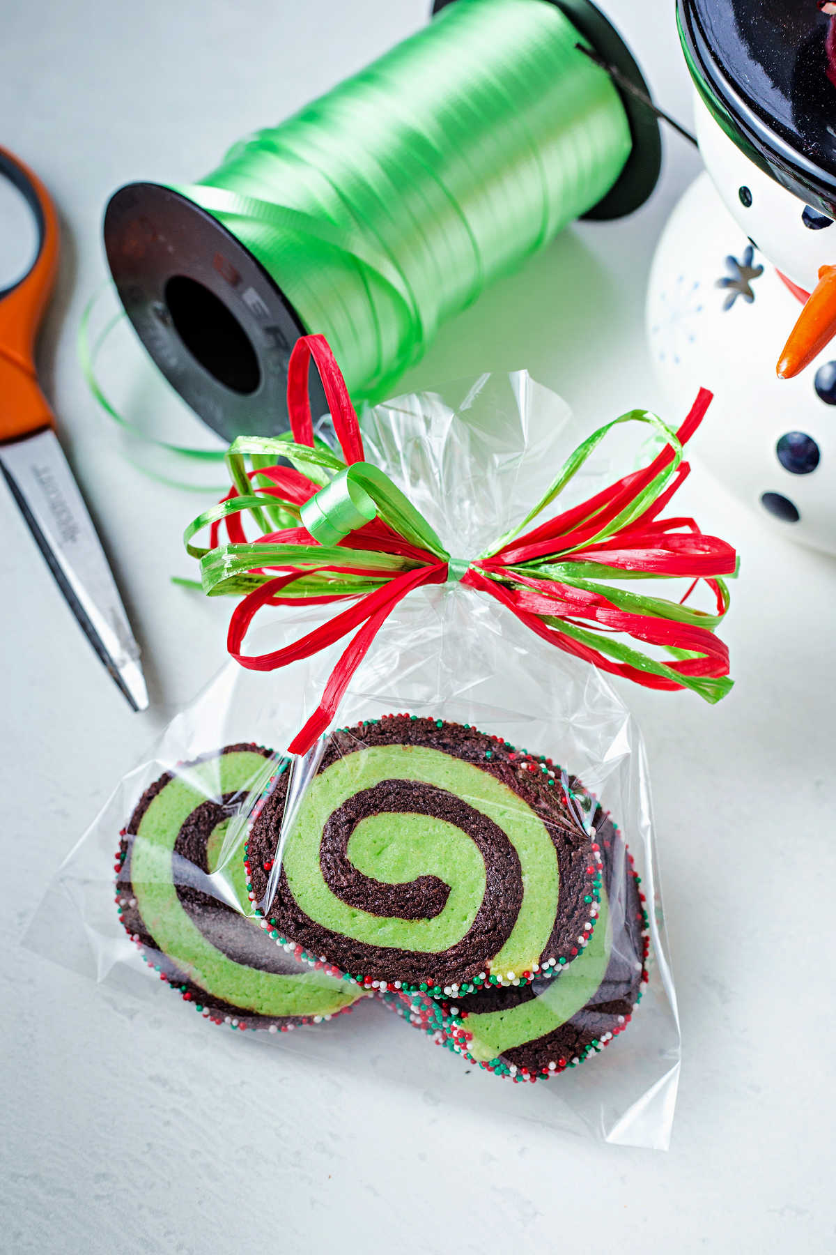 chocolate mint pinwheel cookies packaged in a cellophane bag tied with red and green ribbon on a table with a spool of ribbon and scissors.