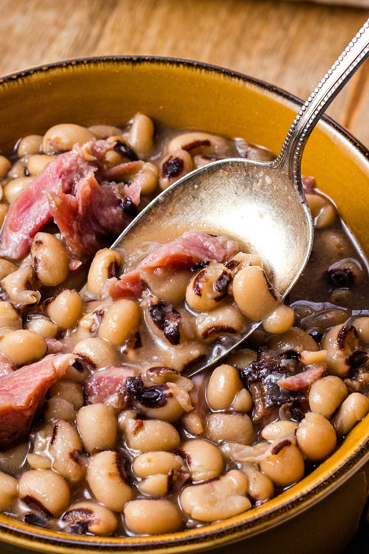 a spoon inserted into a bowl of black eyed peas with chunks of ham.