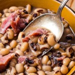 a spoon inserted into a bowl of black eyed peas with chunks of ham.