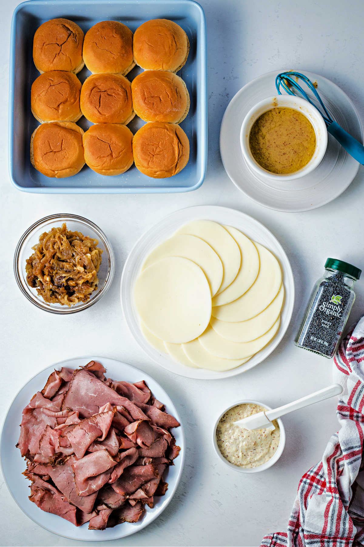 ingredients for making roast beef sliders on a table: slider buns, deli roast beef, horseradish aioli, caramelized onions, provolone cheese slices, and butter sauce.