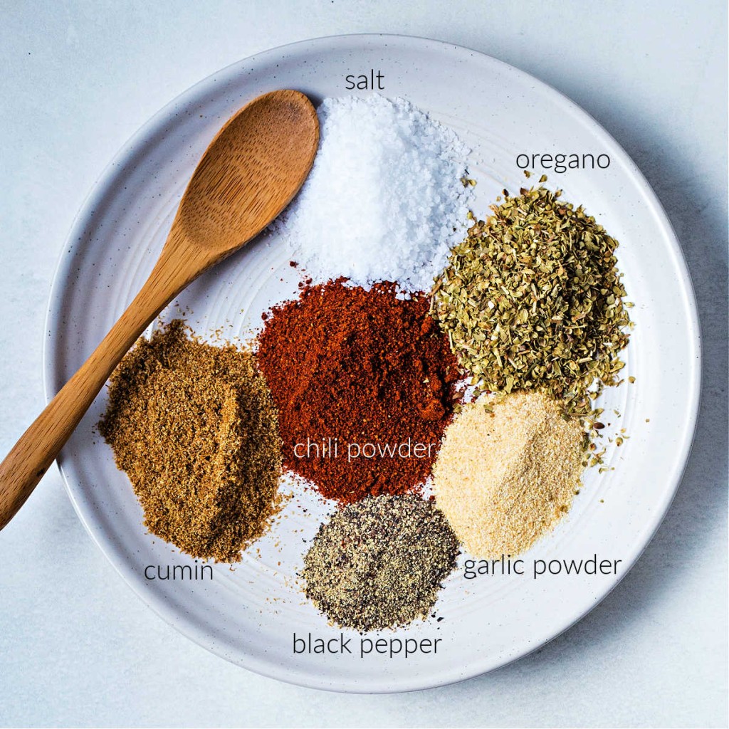 dried spices on a shallow plate with a wooden spoon: salt, oregano, garlic powder, pepper, cumin, and chili powder.