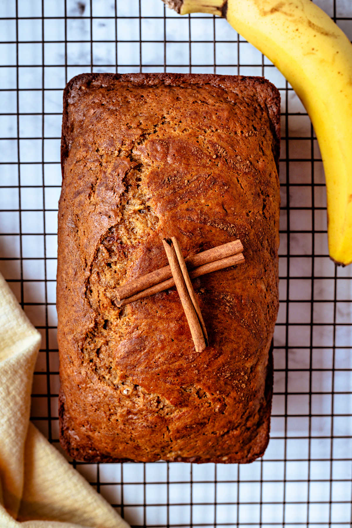 a loaf of whole wheat banana bread on a wire rack with cinnamon sticks on top.