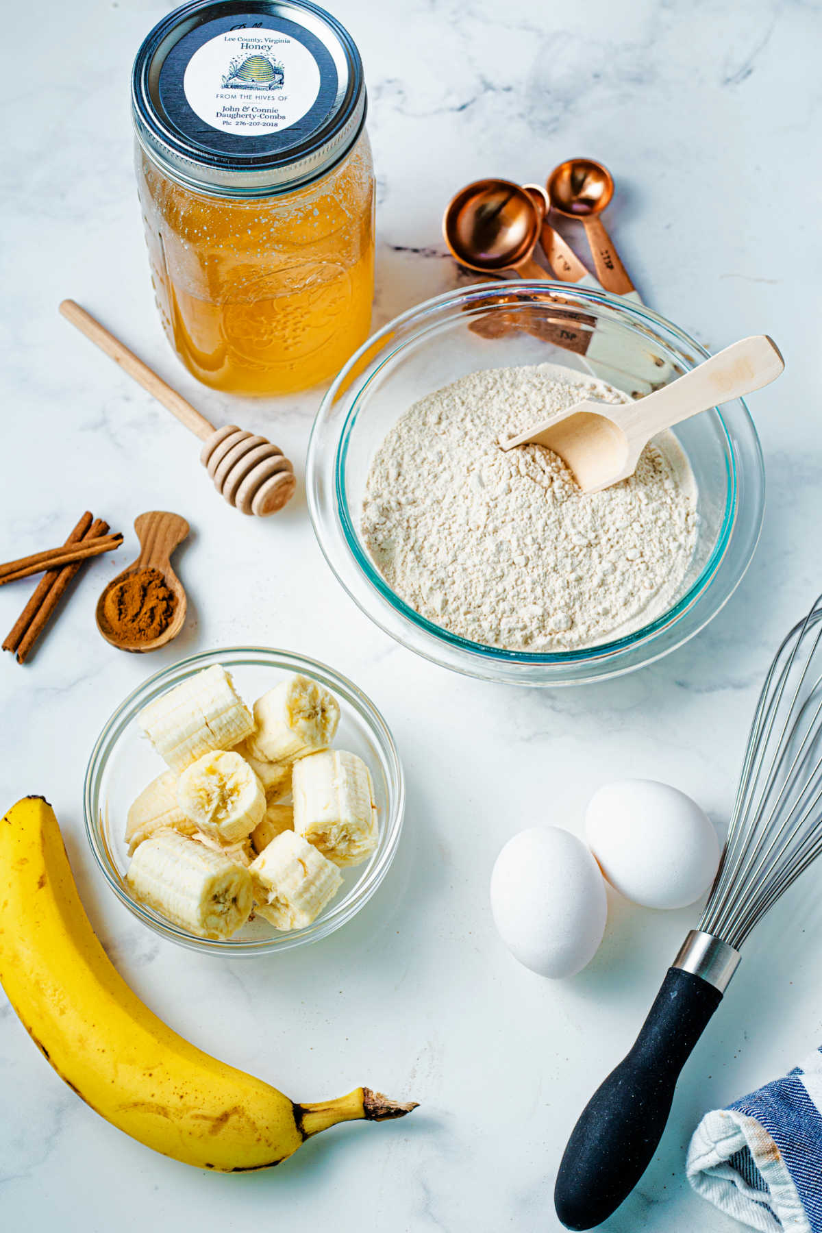 ingredients for whole wheat banana bread on a table, including bananas, honey, eggs, and whole wheat flour.