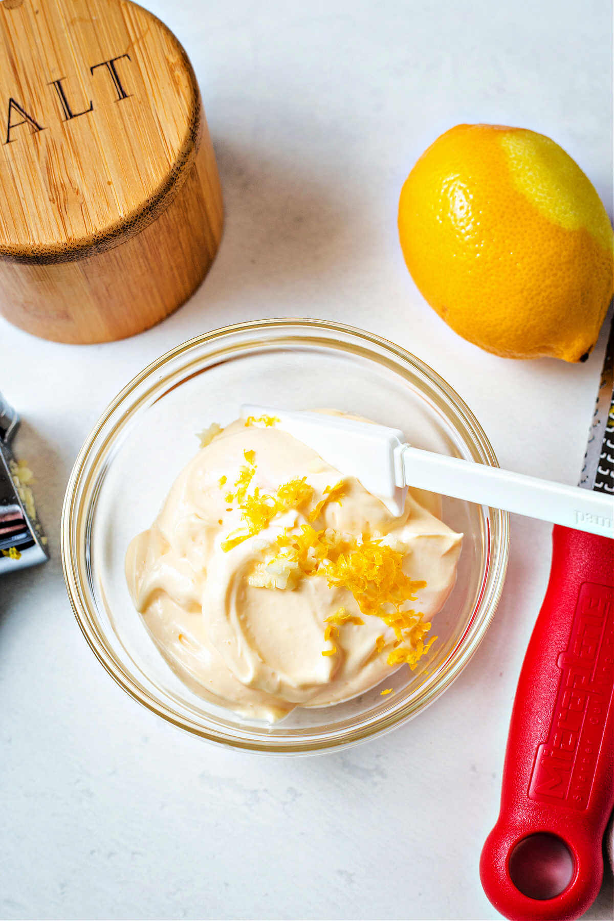 a bowl of mayonnaise with lemon zest on a table with a salt bowl and lemon zester.