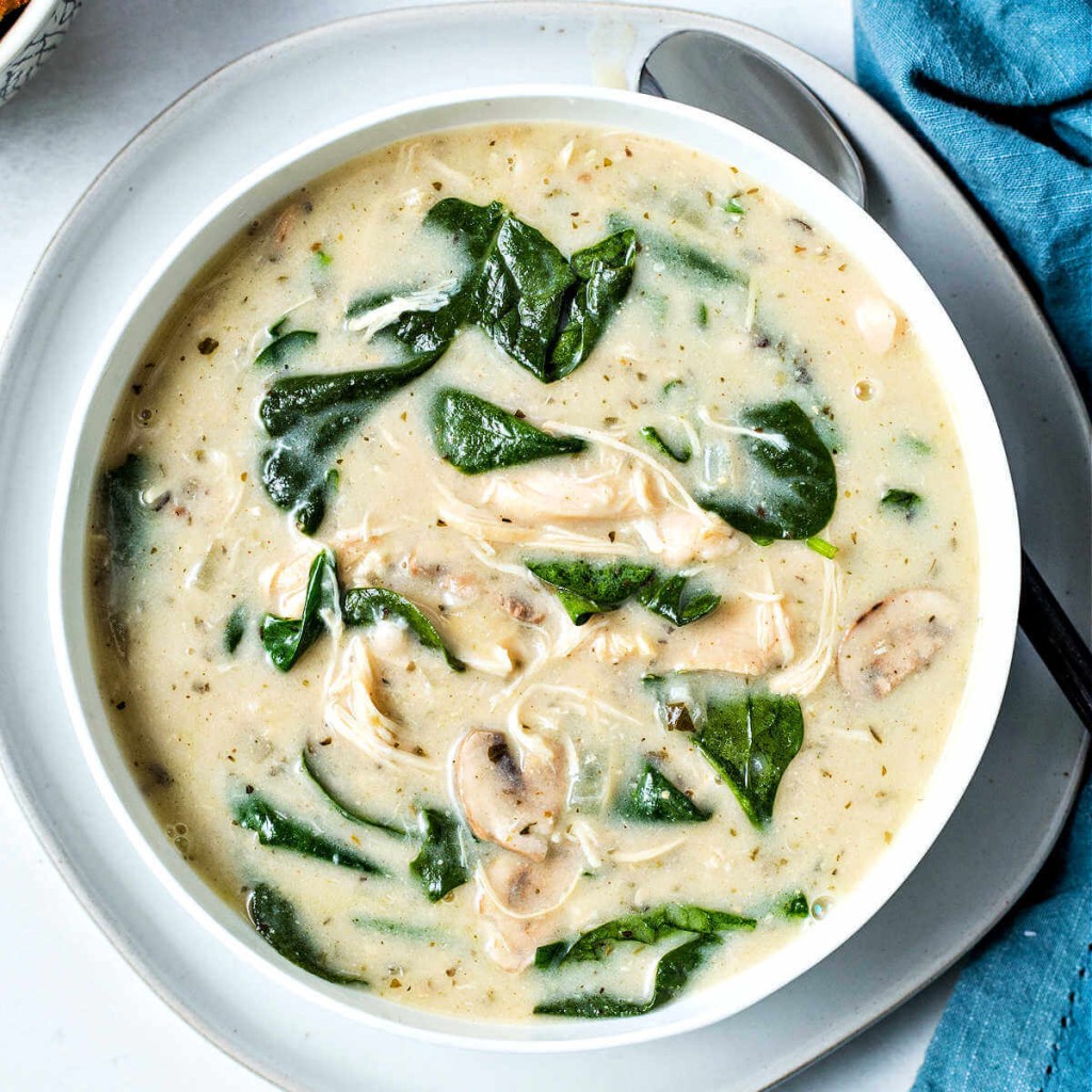 a bowl of chicken florentine soup on top of a plate with a spoon and blue napkin tucked under.