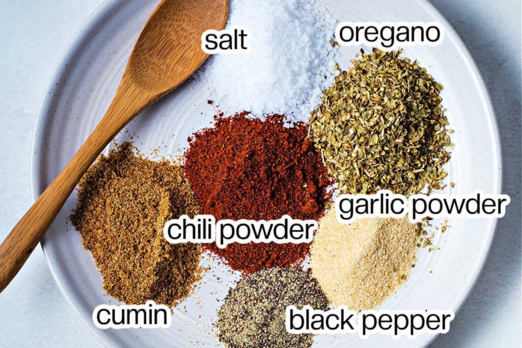 Dried spices on a shallow plate with a wooden spoon: salt, oregano, garlic powder, pepper, cumin, and chili powder.