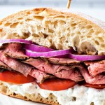 a steak sandwich on a ciabatta bun with tomatoes and red onions.