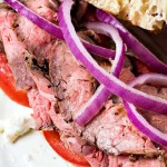 an open face steak sandwich on a ciabatta bun with tomatoes and red onions.