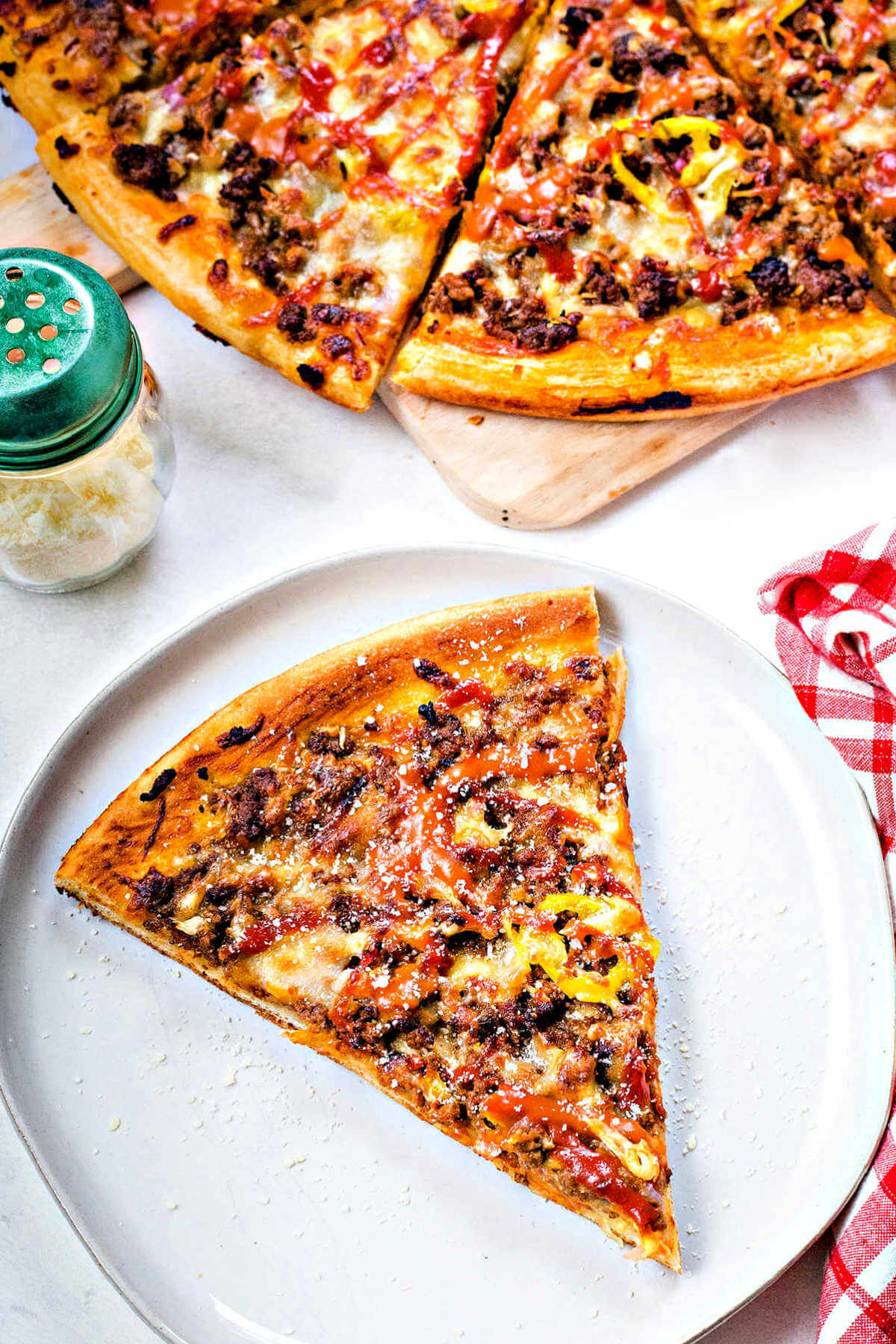 a slice of cheeseburger pizza on a plate with the rest of the pizza in the background.