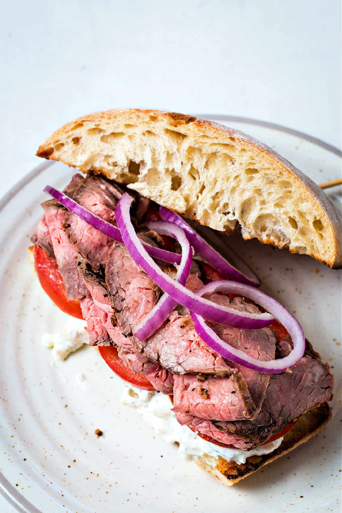 an open face steak sandwich on a ciabatta bun with tomatoes and red onions.