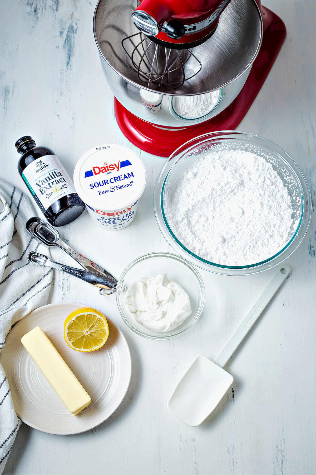 ingredients for sour cream frosting on a kitchen counter: butter, lemon, vanilla, sour cream, and powdered sugar.