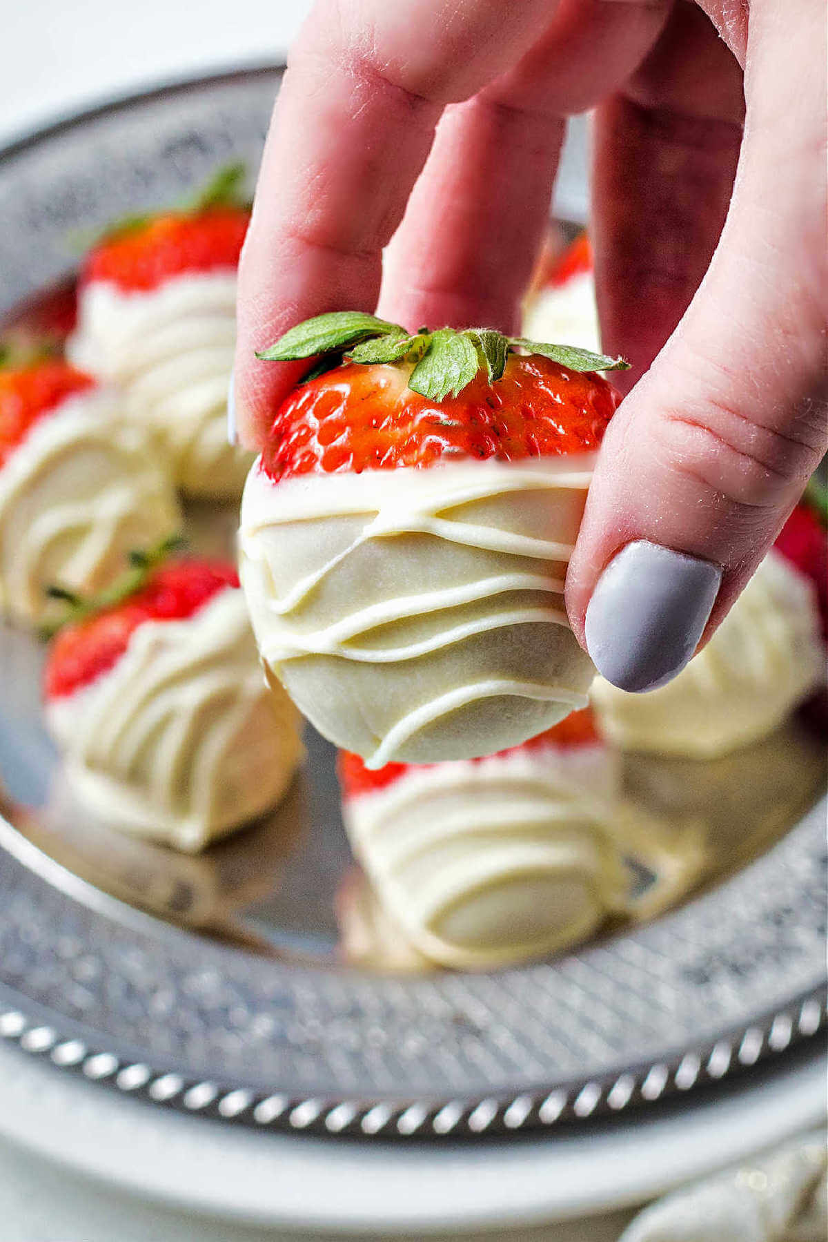 a lady's hand holding a white chocolate dipped strawberry with a plate of strawberries in the background.