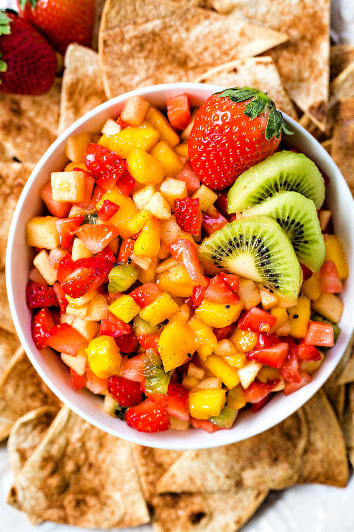 close up of fruit salsa garnished with a whole strawberry in a bowl with cinnamon tortilla chips surrounding it.
