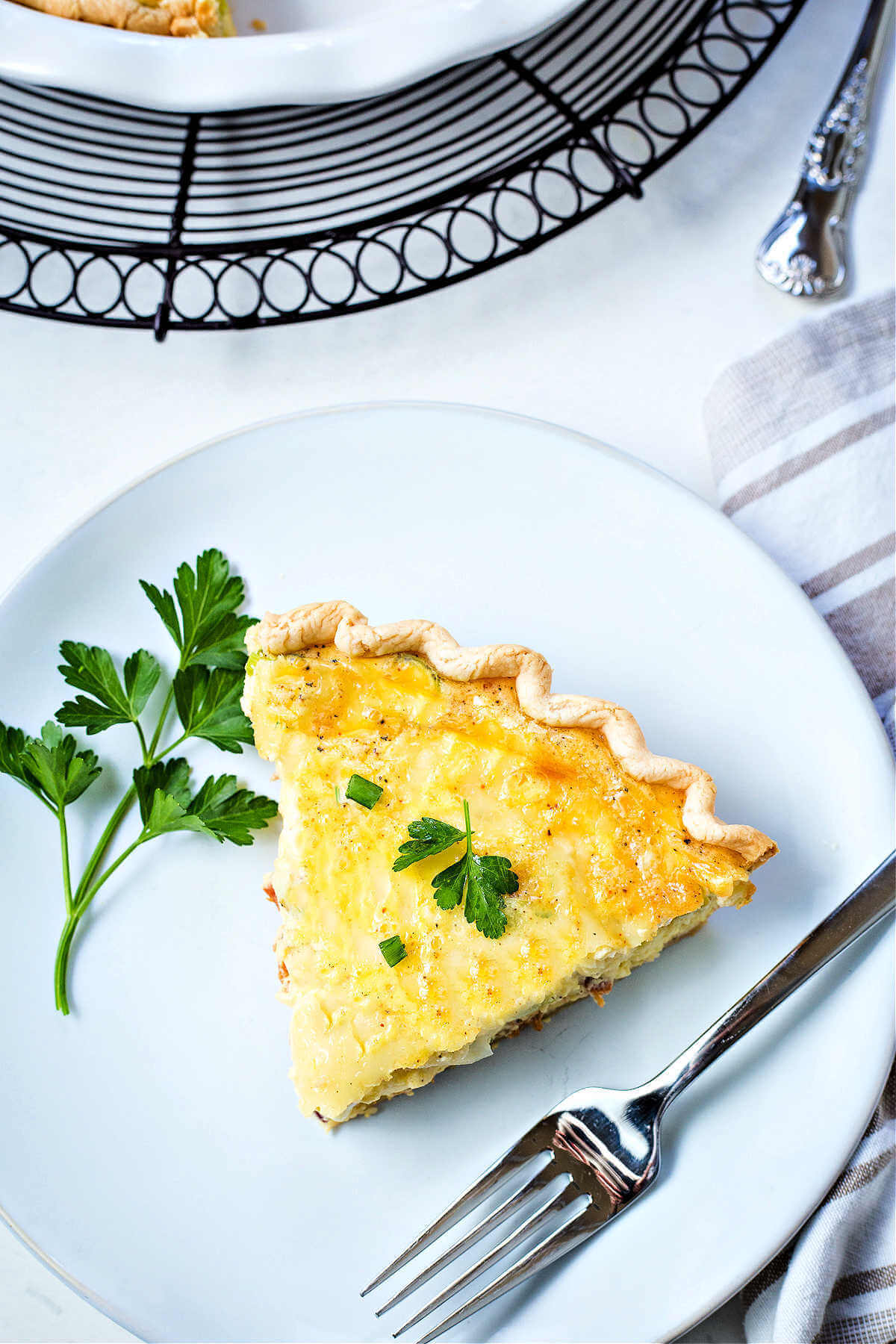 a slice of quiche lorraine on a white plate with a fork garnished with fresh parsley.