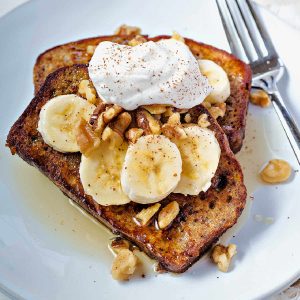 two slices of banana bread french toast on a white plate garnished with sliced bananas, walnuts, and whipped cream with a fork nestled on the plate.