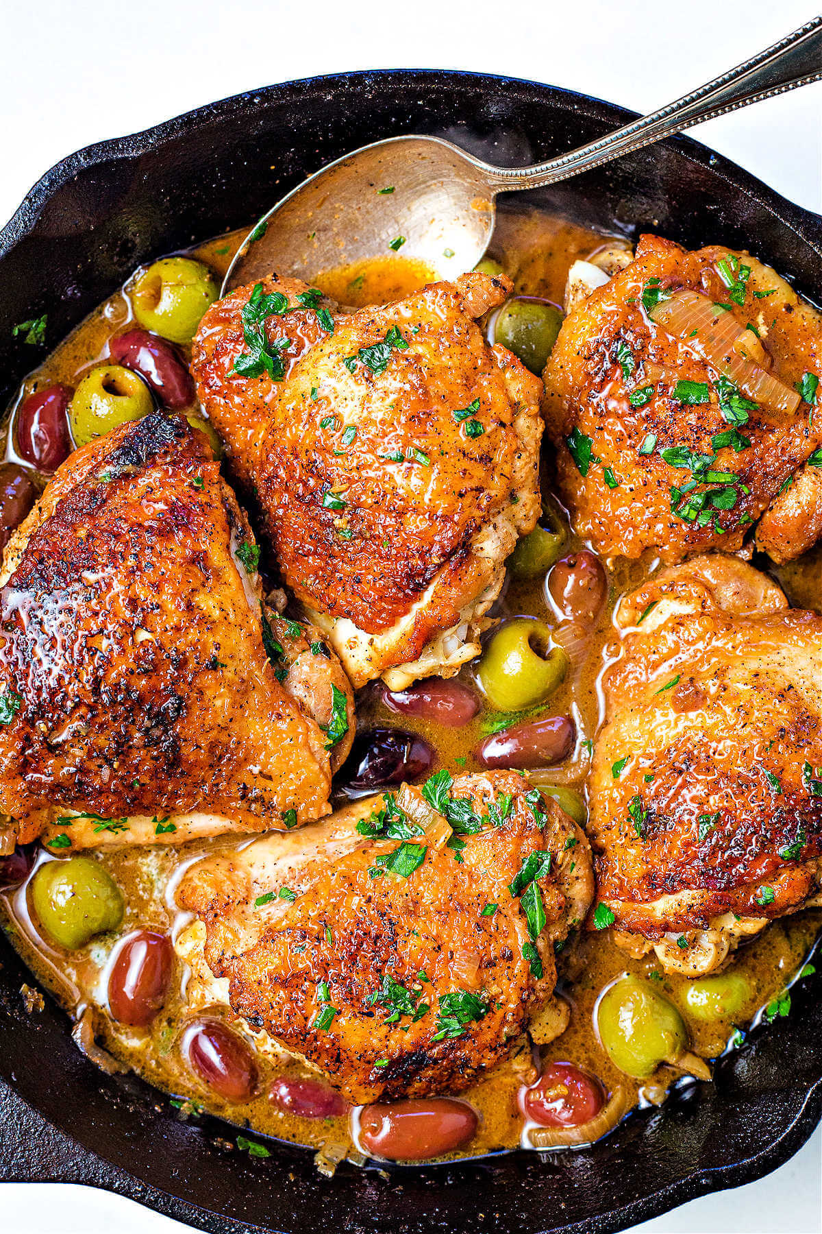 chicken with olives in a cast iron skillet with a spoon drizzling sauce.