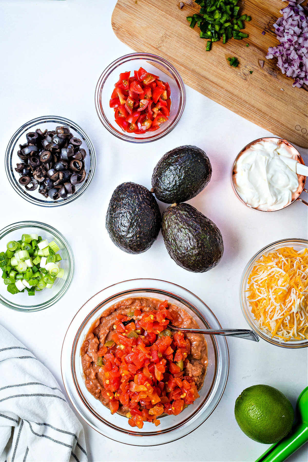 ingredients for layered taco dip on a table, including avocados, olives, tomatoes, cheese, and refried beans.