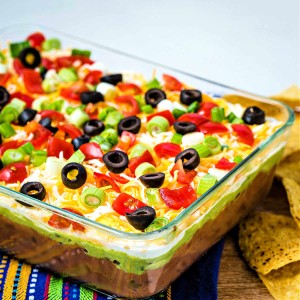 layered taco dip in a glass dish with tortilla chips scattered around.