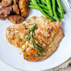 two chicken dijon cutlets on a white plate with green beans and roasted potatoes garnished with rosemary.