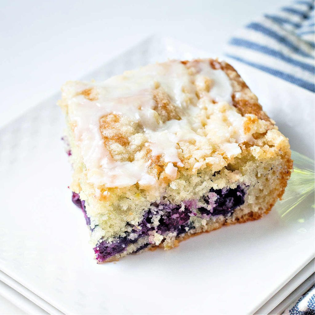 a slice of blueberry crumb cake on a white plate with a blue striped napkin on the table.