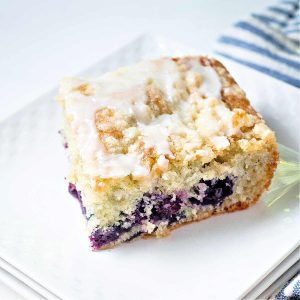 a slice of blueberry crumb cake on a white plate with a blue striped napkin on the table.