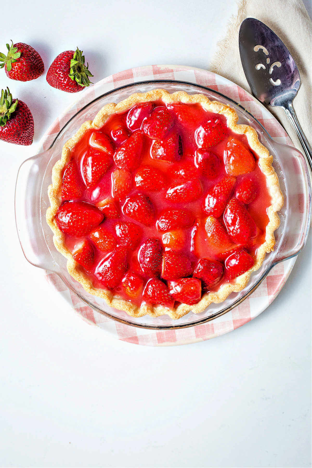 a baked fresh strawberry pie sitting on a pink plate with a pie server and strawberries scattered on a table.