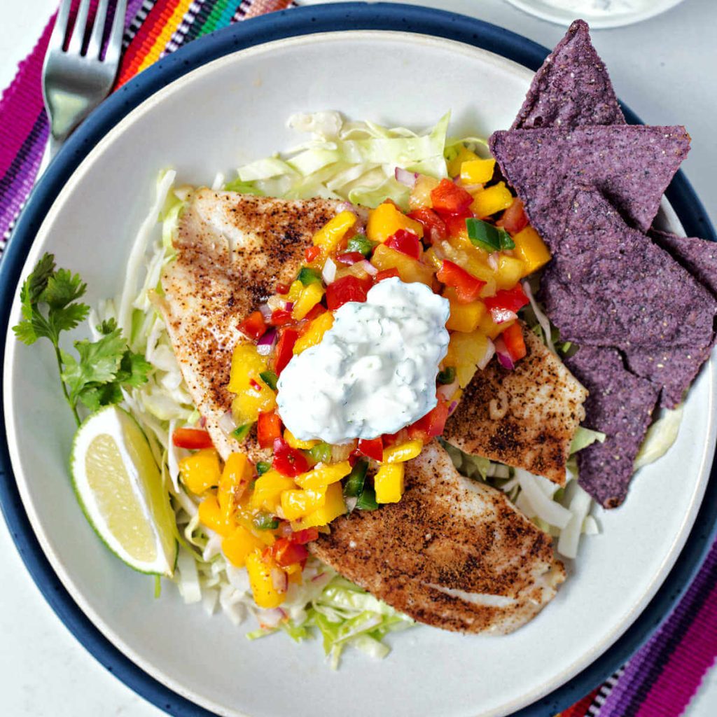 a fish taco bowl garnished with mango salsa and crema with a side of blue tortilla chips and a slice of lime.