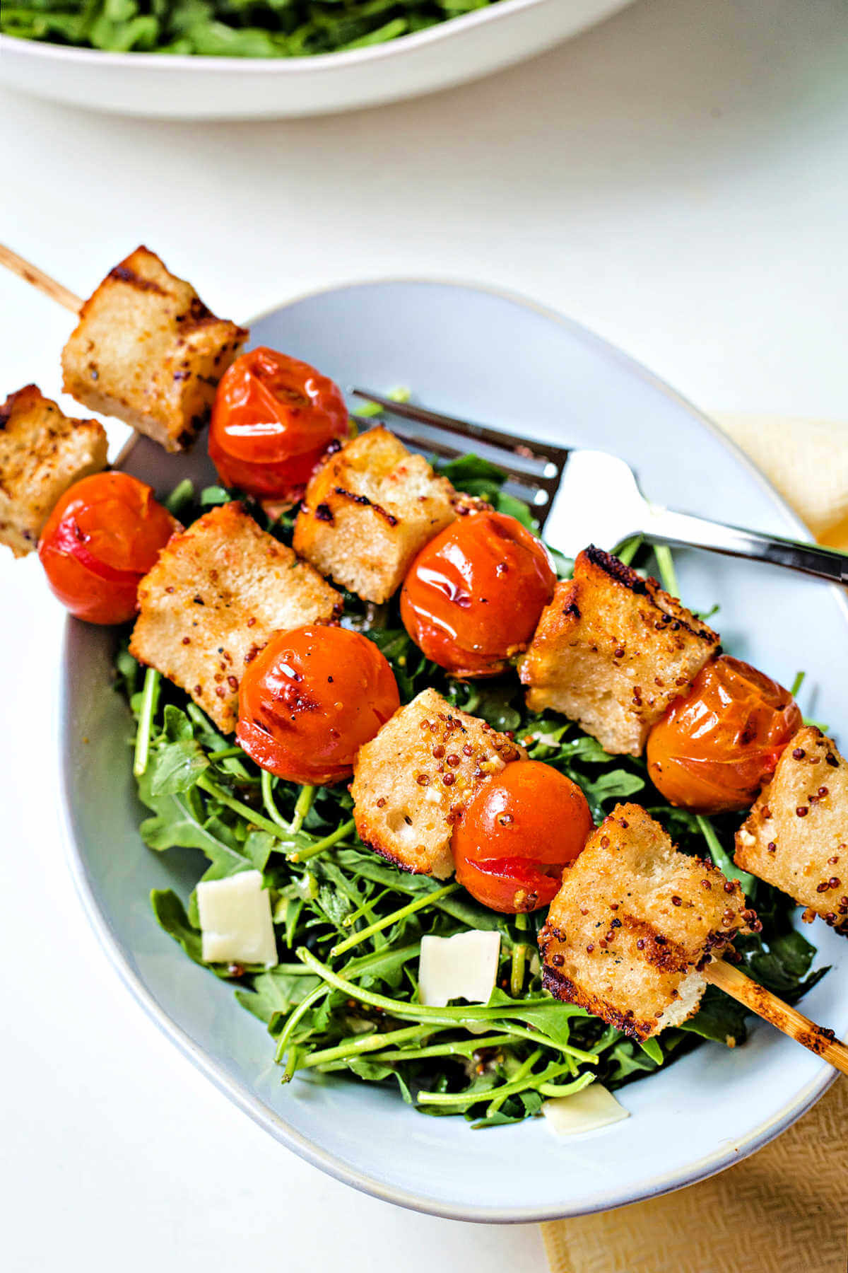 skewers of bread and tomatoes laying on top of a bowl of arugula with shaved parmesan for panzanella salad.