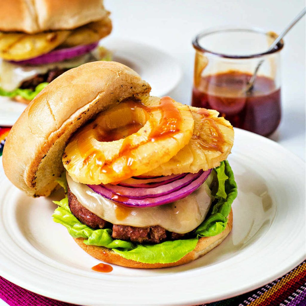 an open face hawaiian burger with provolone cheese, grilled pineapple, and red onion and drizzled with sauce on a plate.