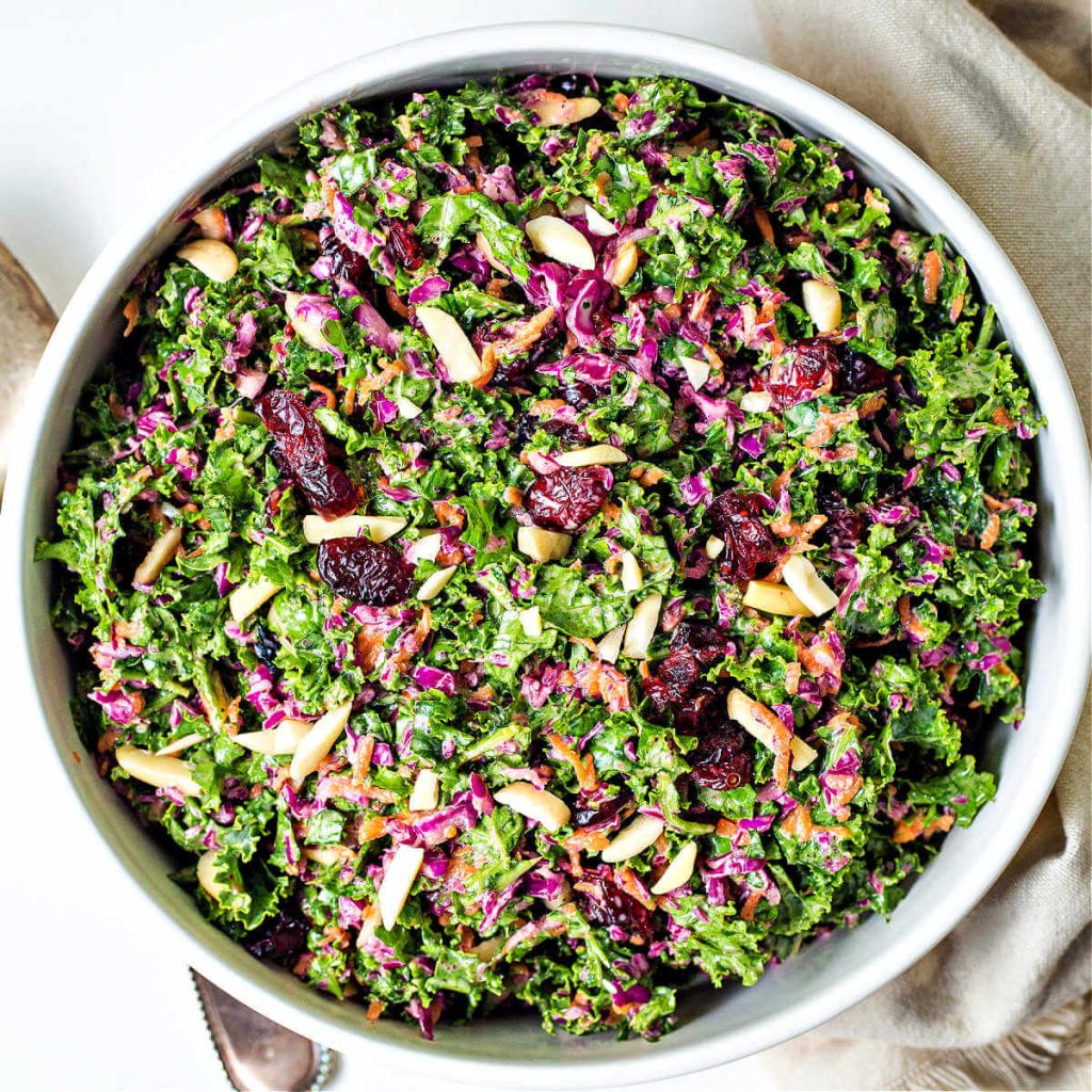 kale slaw with purple cabbage, carrots, dried cranberries, and slivered almonds in a white bowl on a table.