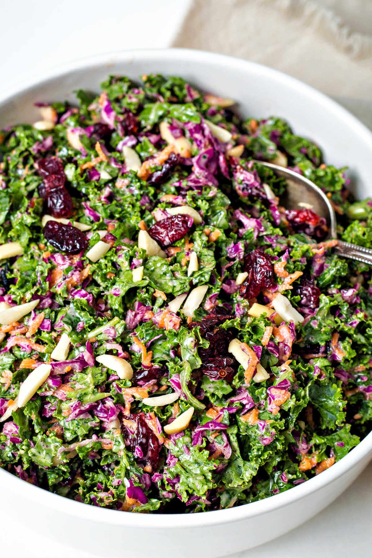 kale slaw with purple cabbage, carrots, dried cranberries, and slivered almonds in a white bowl with a serving spoon inserted sitting on a table.
