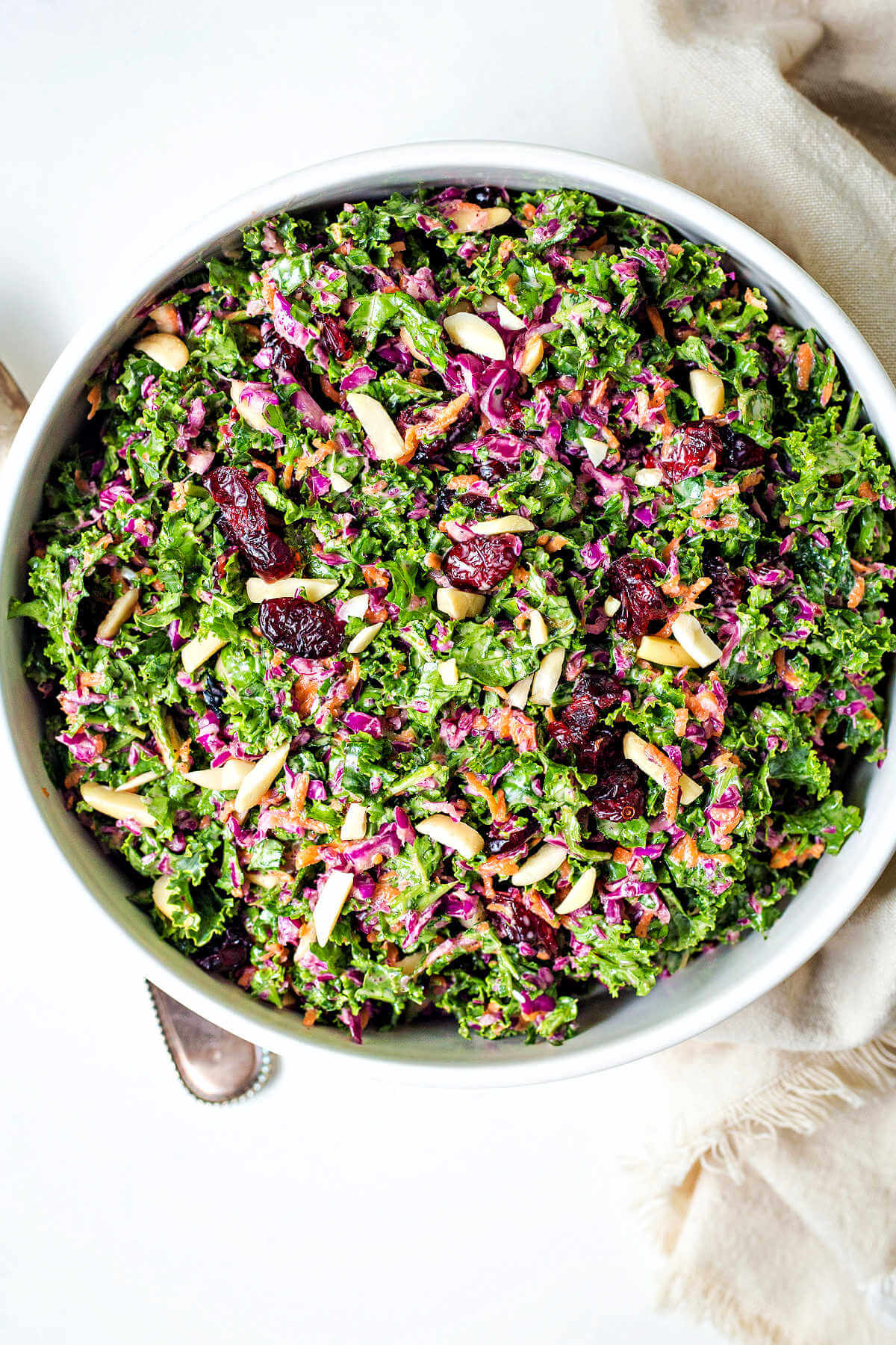 kale slaw with purple cabbage, carrots, dried cranberries, and slivered almonds in a white bowl on a table.
