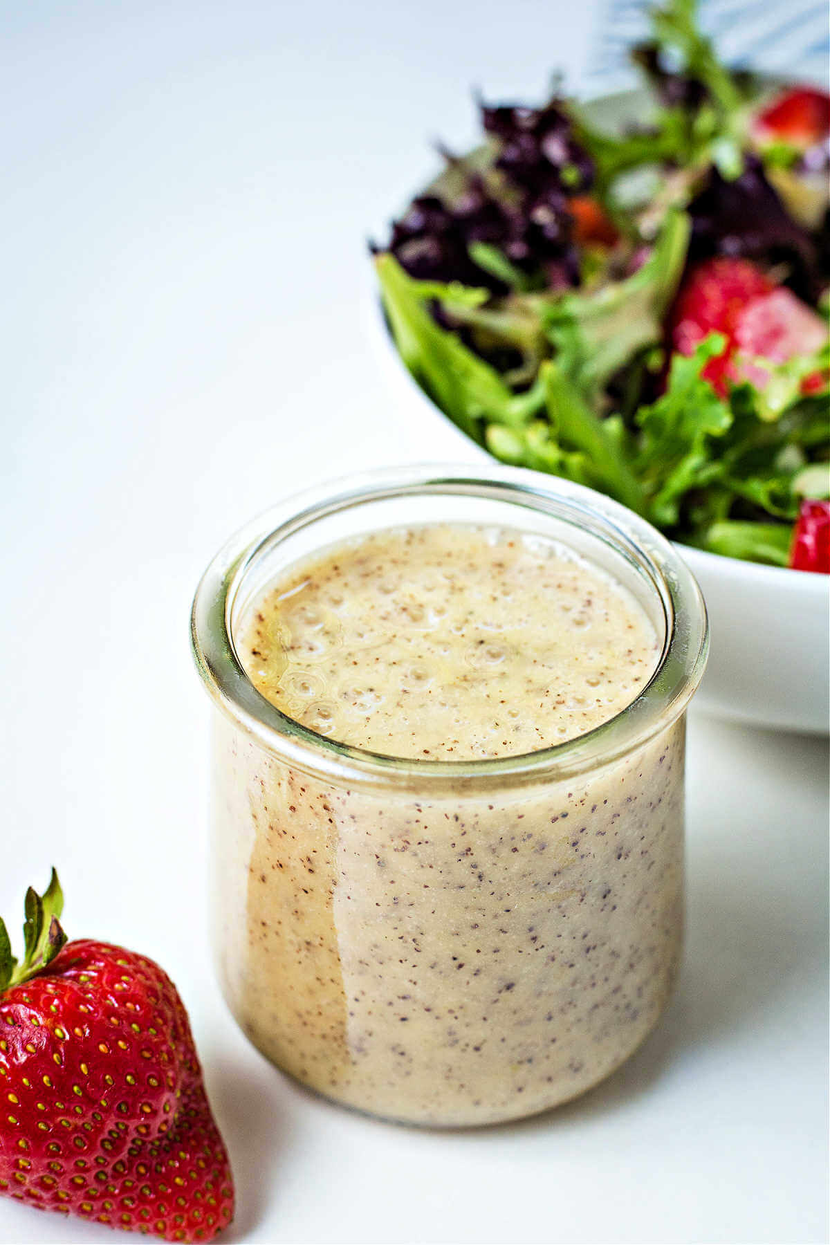 poppy seed dressing in a small glass jar sitting on a table in front of a bowl of salad.