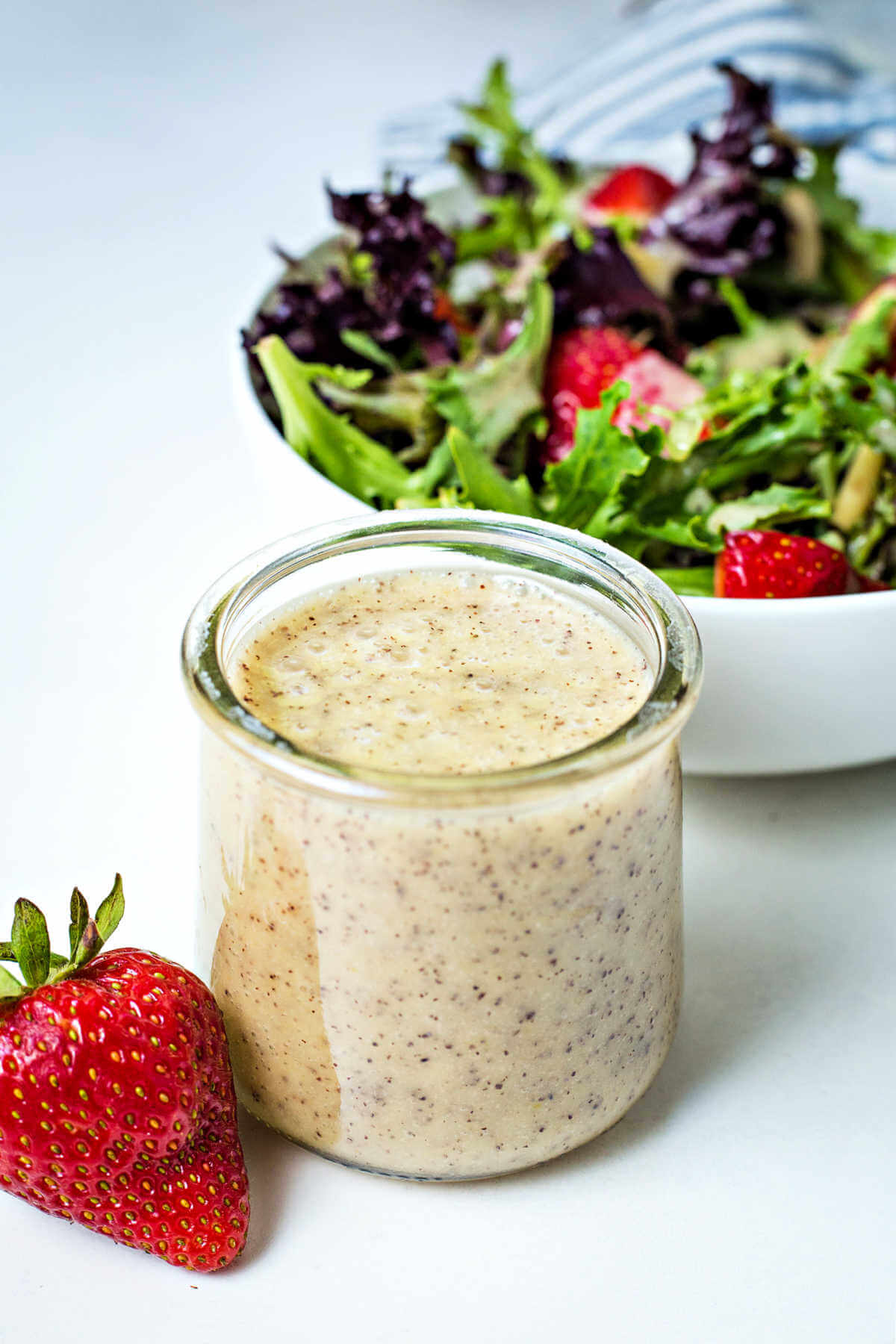 poppy seed dressing in a small glass jar sitting on a table in front of a bowl of salad.