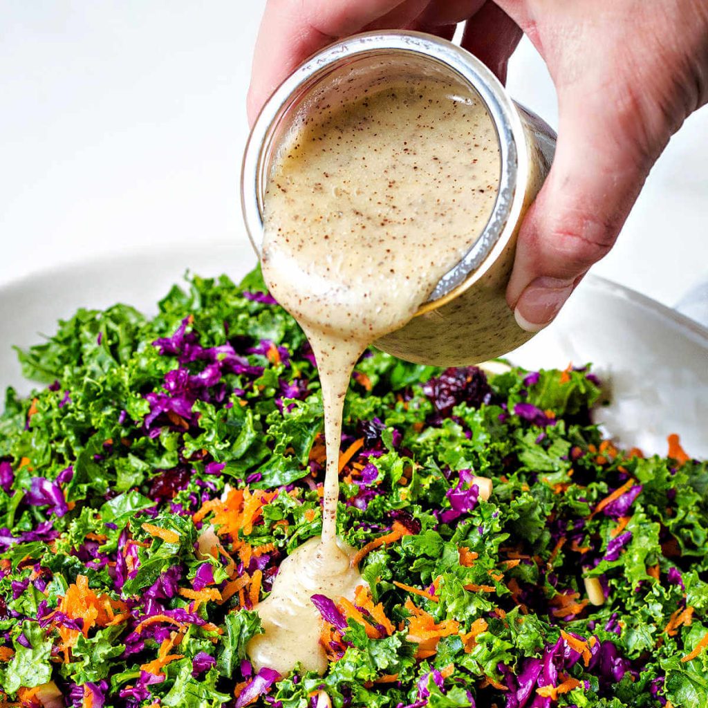 pouring poppy seed dressing over a bowl of kale salad.