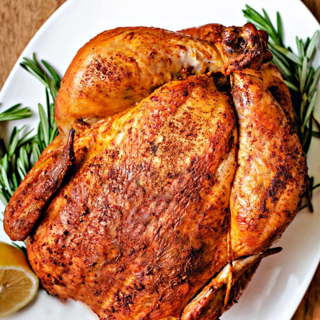 a smoked whole chicken on a white platter with lemon half and rosemary sprigs.