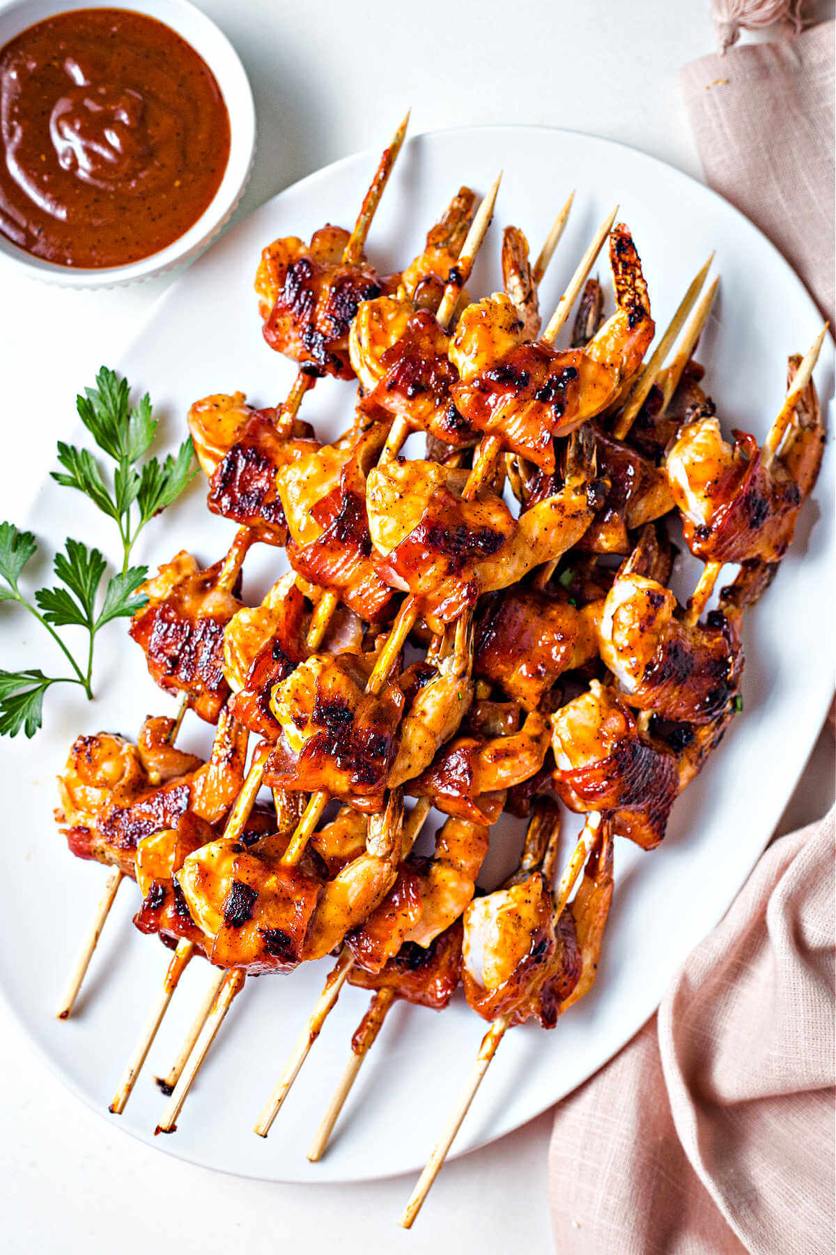 a platter filled with skewers of bacon wrapped shrimp with a bowl of barbecue sauce to the side.