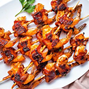a platter filled with skewers of bacon wrapped shrimp with a bowl of barbecue sauce to the side.